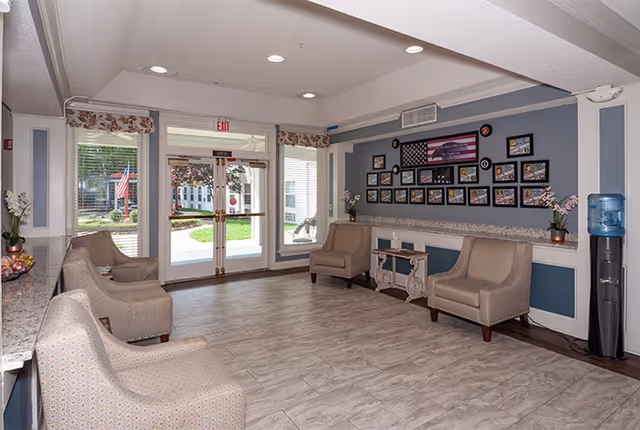 A bright and clean assisted living community lobby with beige armchairs arranged along the walls, a water cooler on the right, and a wall decorated with framed pictures and an American flag. Double glass doors lead outside to a green lawn with a flagpole visible through the windows.