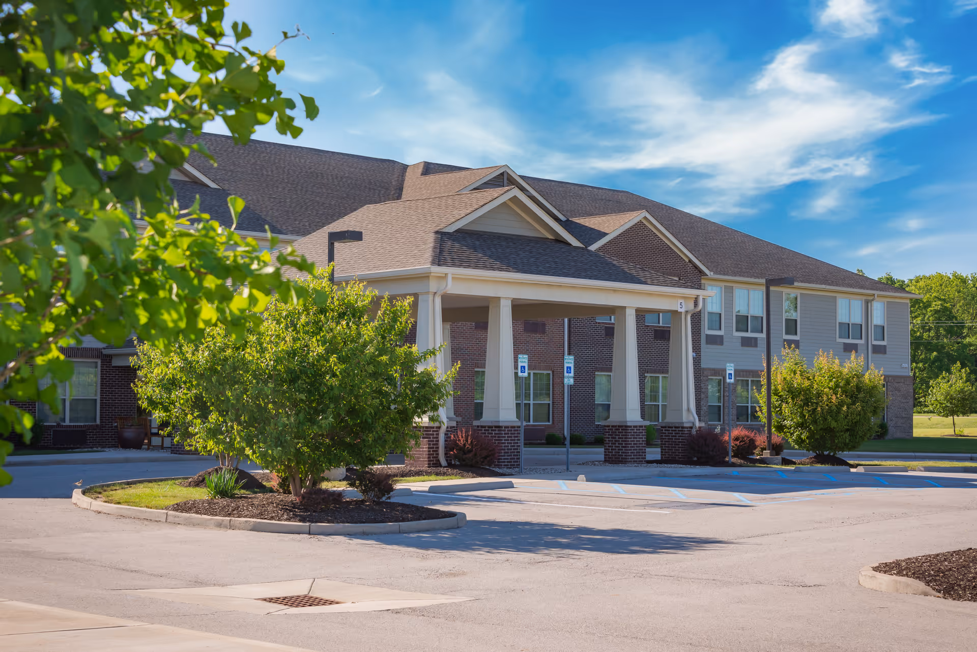Exterior view of a senior living facility building with a covered entrance, surrounded by greenery and a parking lot with handicap parking spaces under a partly cloudy blue sky.