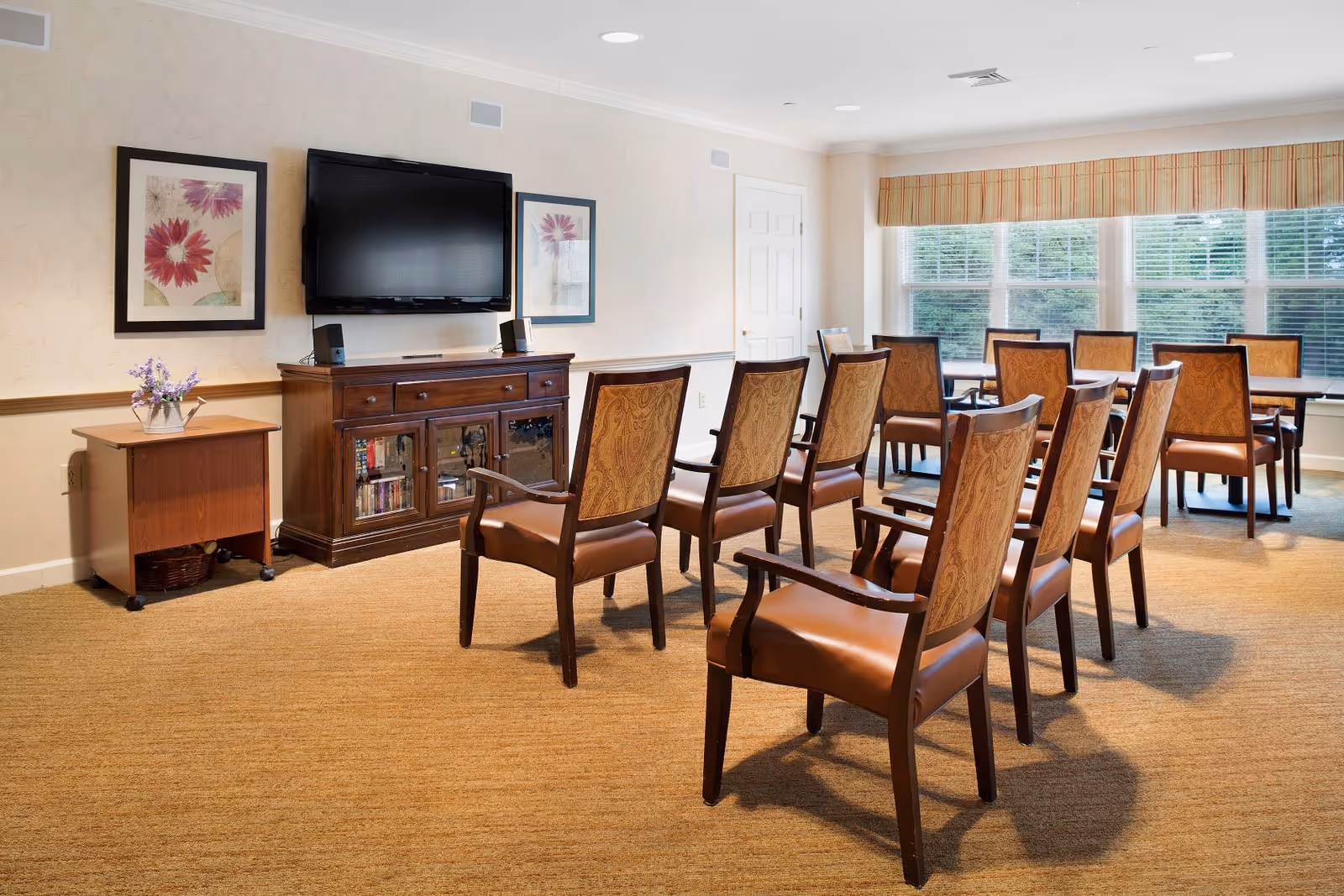 A well-lit room with multiple wooden chairs arranged in rows facing a wall-mounted flat screen TV above a wooden cabinet. The room has beige walls, carpeted floor, two framed floral pictures on the wall, a small side table with a vase of flowers, and large windows with blinds and a striped valance letting in natural light.