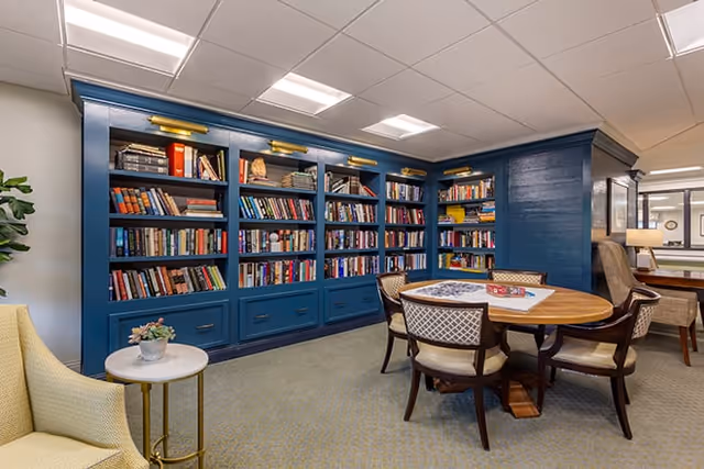 A cozy reading and game area in a senior living facility featuring a large blue bookshelf filled with books, a round wooden table with four chairs, and a puzzle and board game on the table. There is a small side table with a plant and comfortable seating in the background.