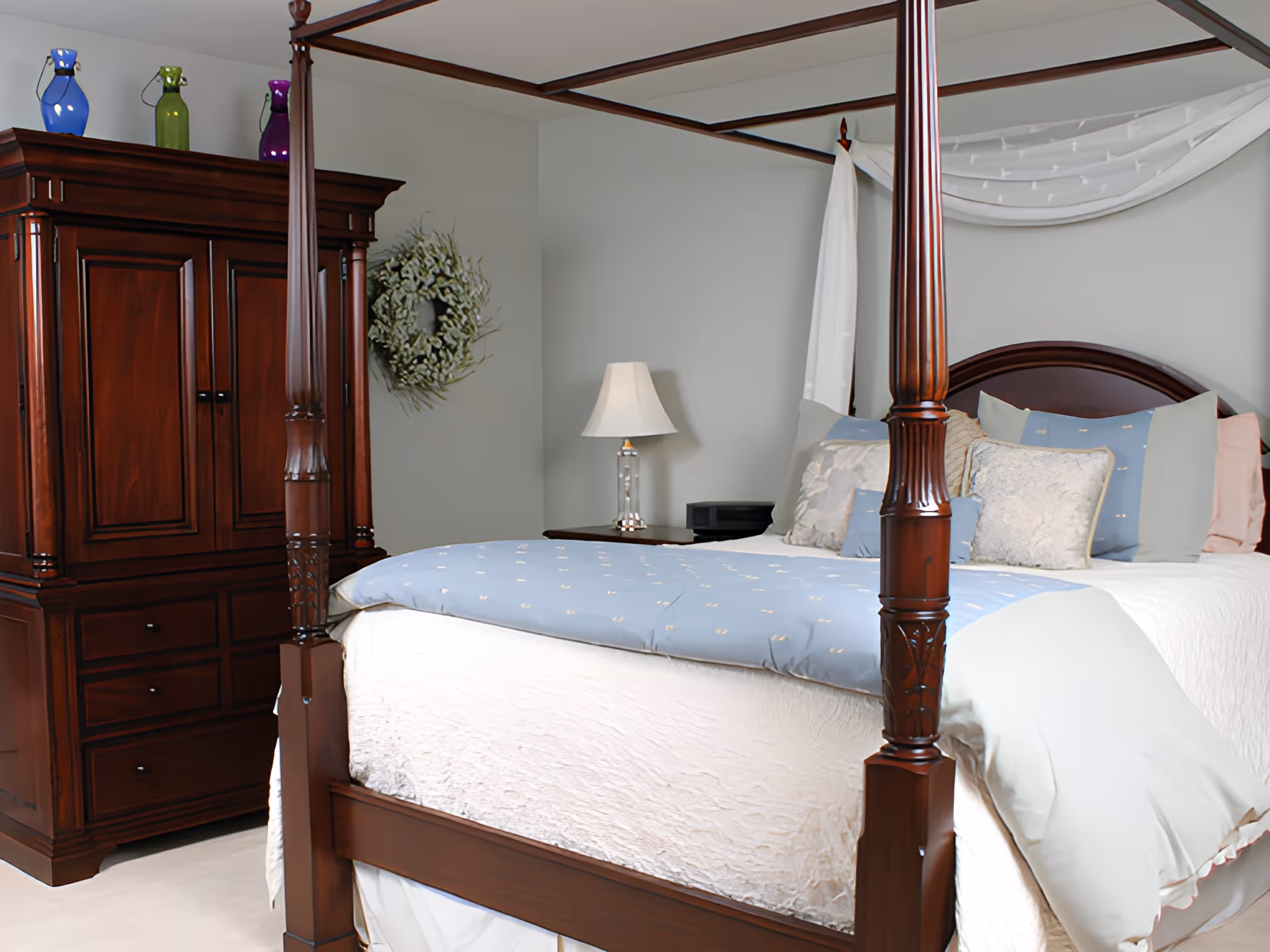 A bedroom with a large wooden four-poster bed adorned with multiple pillows and a light blue comforter. Next to the bed is a wooden nightstand with a lamp and a small black device. On the left side of the room is a wooden armoire with colorful decorative vases on top. A green wreath hangs on the light gray wall behind the bed.