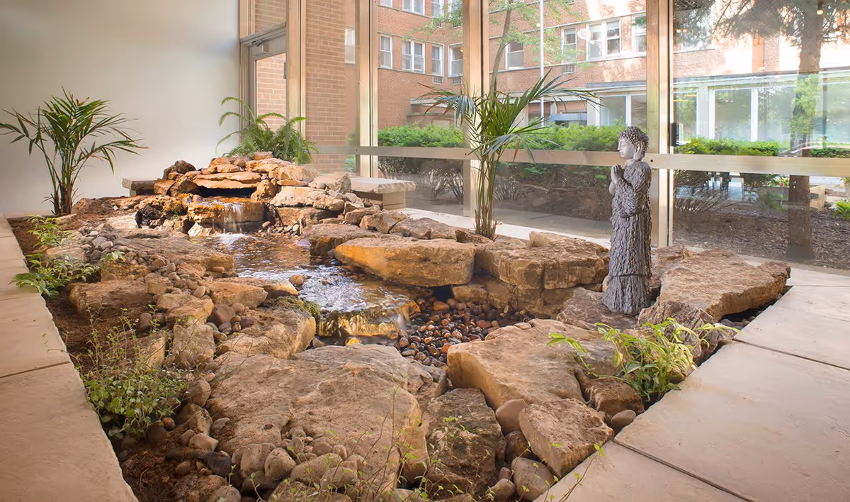 Indoor water feature with rocks, small plants, and a statue of a praying figure, located in a sunlit area with large windows showing an exterior courtyard and brick building.