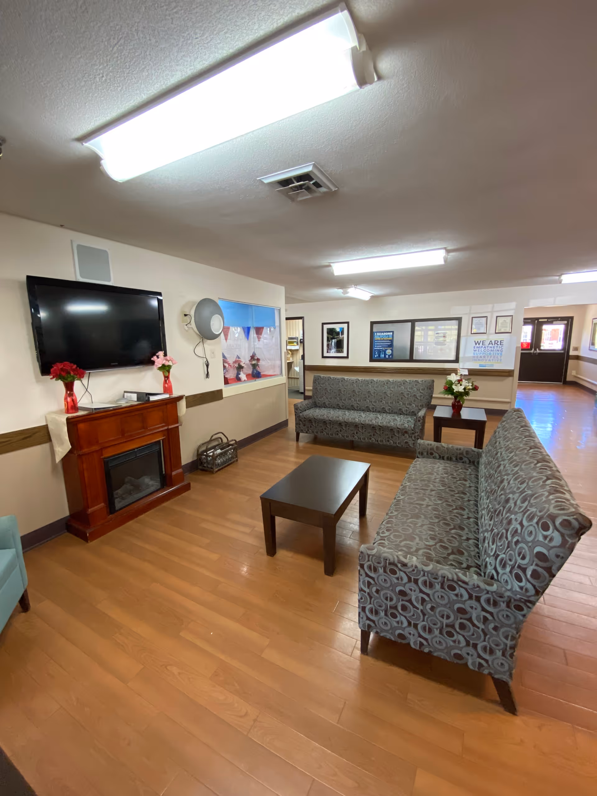 A cozy common area with two patterned sofas facing each other across a dark wooden coffee table. A flat-screen TV is mounted on the wall above a wooden electric fireplace with two red vases holding flowers on top. The room has wood flooring, bright overhead lighting, and framed pictures and notices on the walls. There is a window opening to another room and a hallway leading to an exit door.