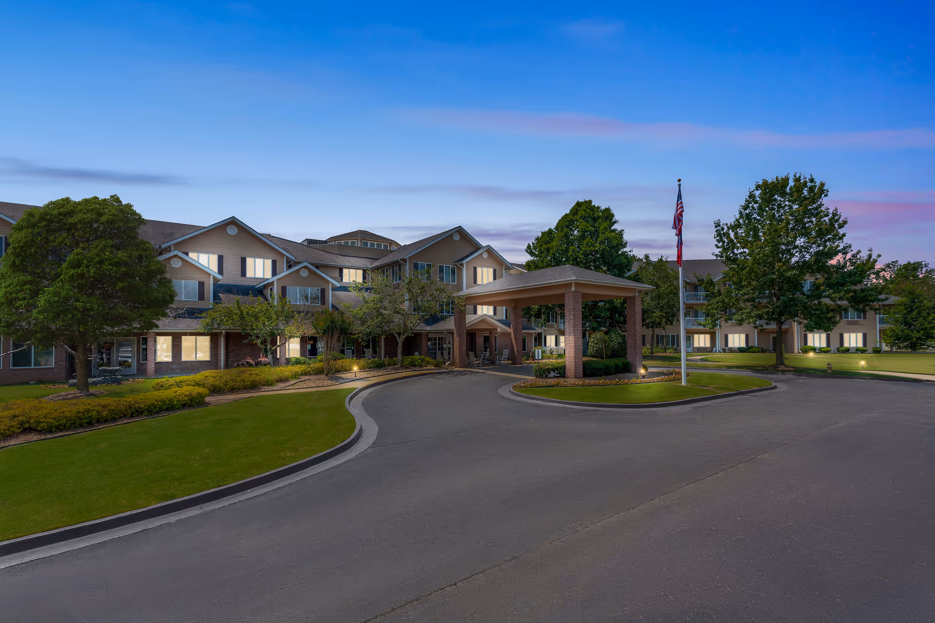 Front exterior of a large senior living facility at dusk with a covered drive-up entrance, landscaped lawns, trees, and an American flag.