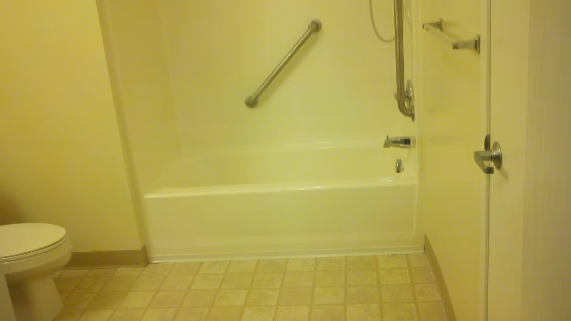 A bathroom with a white bathtub featuring a metal grab bar, a toilet to the left, and a door with a metal handle on the right. The floor has a beige and light brown tiled pattern.