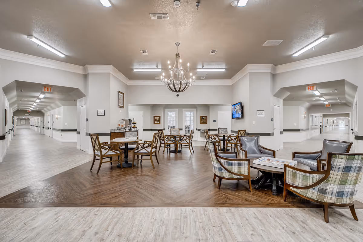 A spacious common area in a senior living facility featuring a chandelier, several tables with chairs, and a seating area with armchairs around a coffee table. The room has light-colored walls and flooring with a darker wood section in the center. Hallways extend on both sides with exit signs visible.