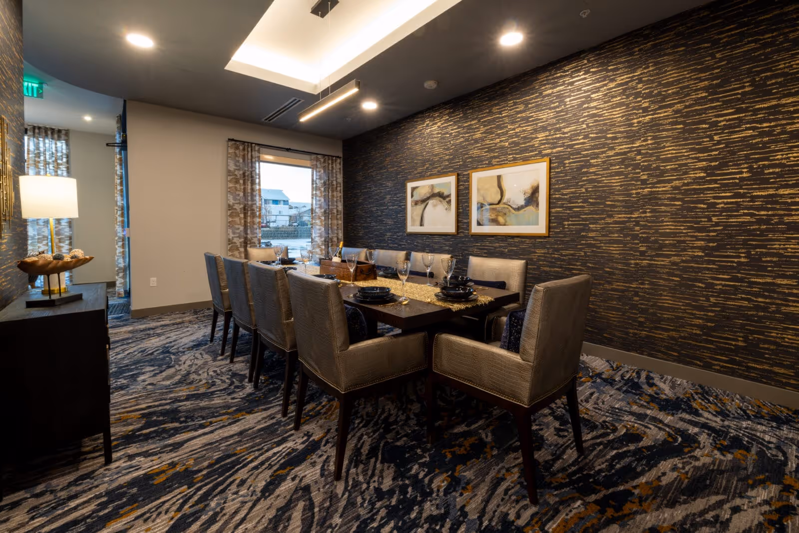 A modern dining room with a long rectangular table set for eight people. The chairs are upholstered in a light gray fabric with dark wooden legs. The table is decorated with black plates, wine glasses, and a centerpiece. The room features a patterned carpet with blue, gray, and gold tones, a dark accent wall with gold streaks, and two abstract framed artworks. A window with patterned curtains allows natural light into the room, and a sideboard with a lamp and decorative bowl is visible on the left.