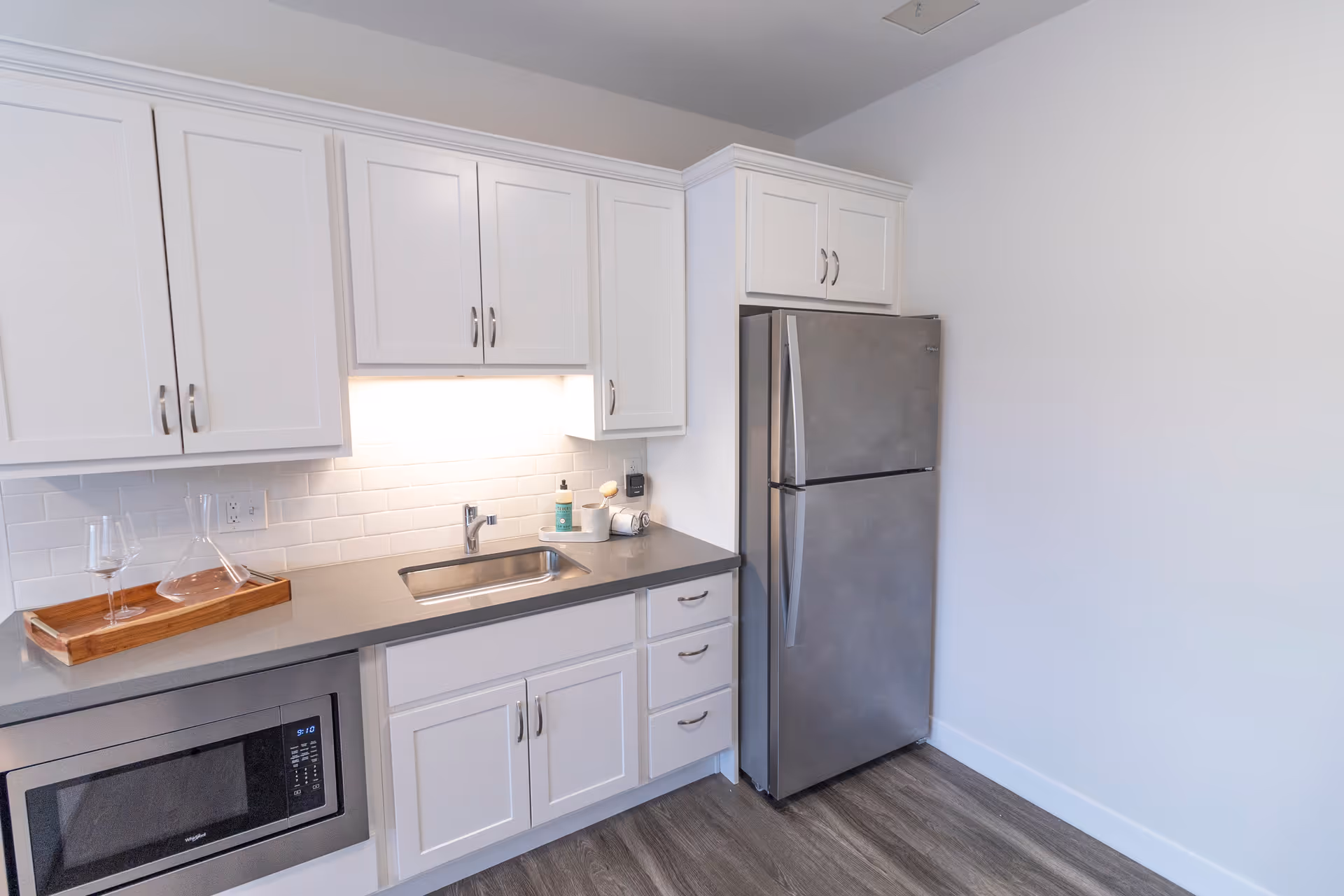 A modern kitchen area with white cabinets, a stainless steel refrigerator, a built-in microwave, a sink with a faucet, and a gray countertop. There is a wooden tray with two wine glasses and a decanter on the counter. The backsplash is white subway tile, and the floor has a wood-like finish.