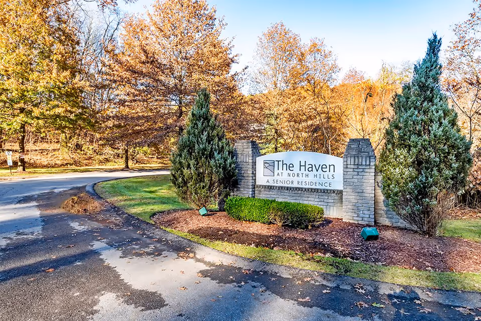 Entrance sign for The Haven at North Hills Senior Residence surrounded by landscaped bushes and trees with autumn foliage, next to a curved paved road.