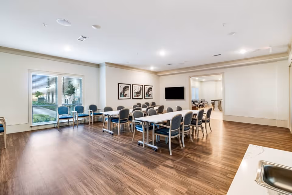 Bright multipurpose dining/activity room with tables and chairs, a wall-mounted TV, and large windows.