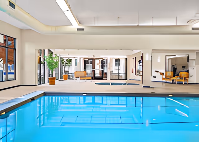 Indoor swimming pool area with clear blue water, surrounded by beige tiled flooring and large windows. There are potted plants and seating areas with chairs and tables visible in the background.