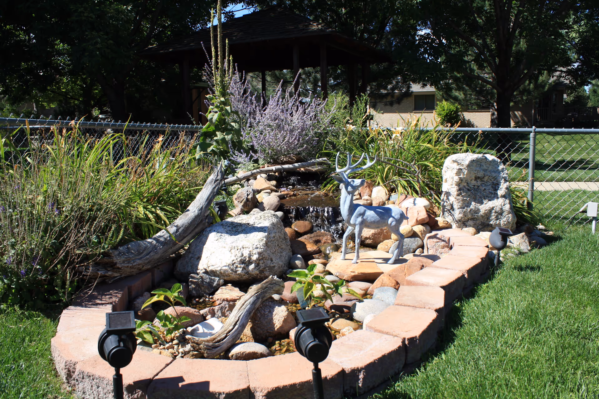 A landscaped garden area with a small water feature surrounded by rocks and plants. There is a decorative metal deer statue placed on a stone platform in the center. The garden is bordered by a low brick wall and has solar-powered garden lights. In the background, there is a chain-link fence, trees, and a gazebo.