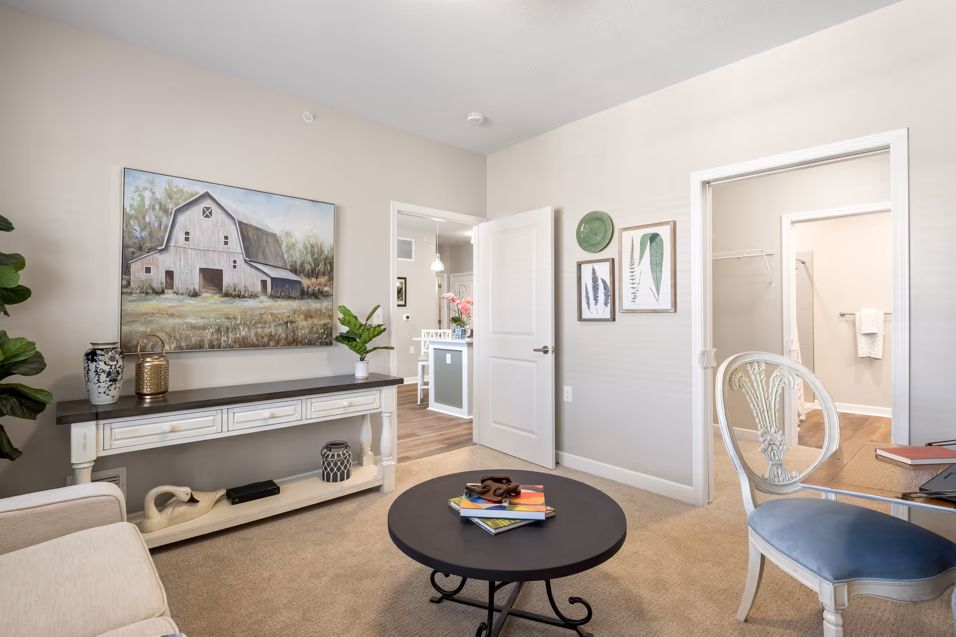 A cozy living room area with a beige couch, a round black coffee table with books and decorative items, and a white console table against the wall with vases and a plant. A large painting of a barn hangs above the console table. The room has beige walls and carpet, with an open door leading to a kitchen area and another open doorway showing a bathroom and a chair with a blue cushion next to a small table.