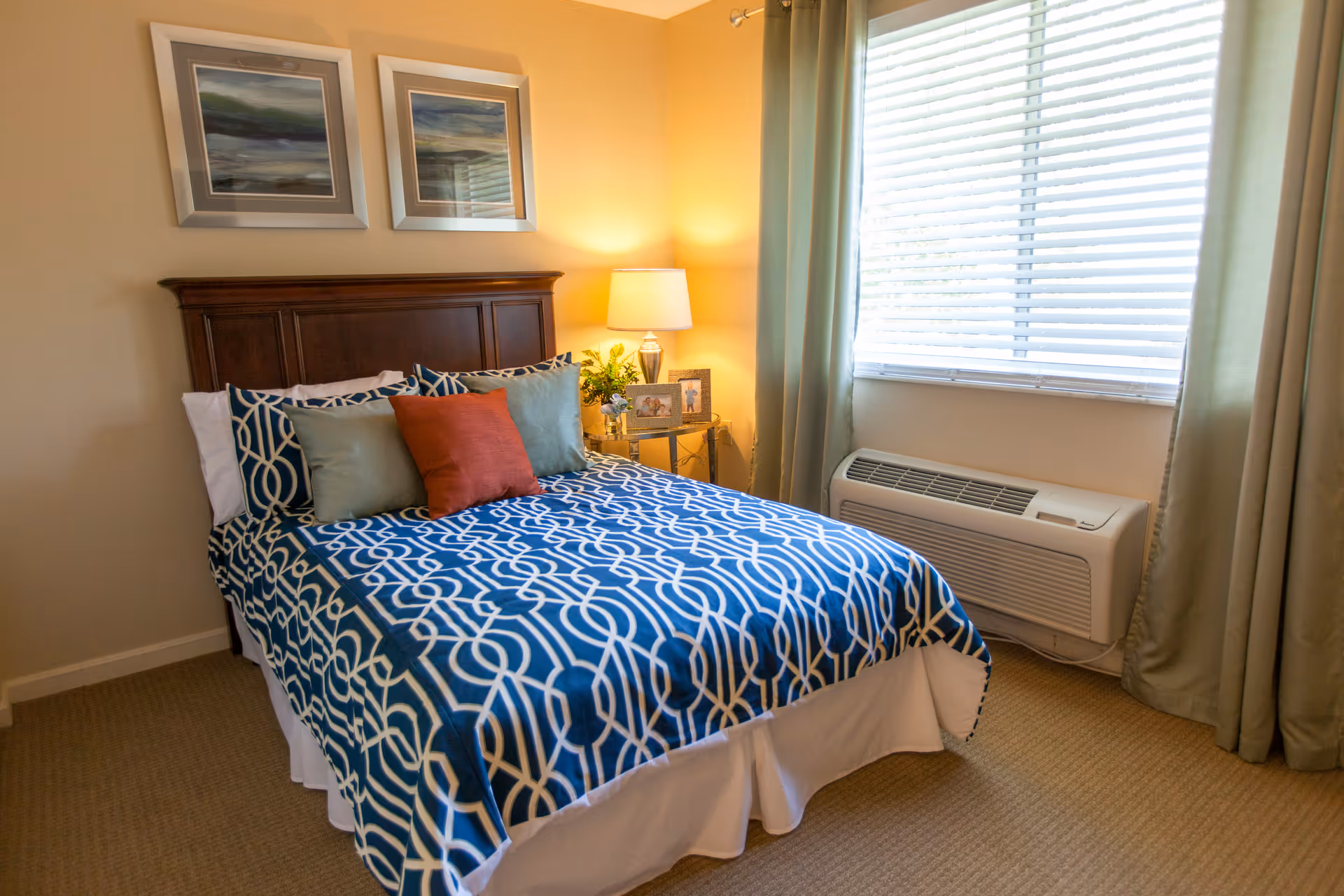 A neatly made bedroom featuring a blue patterned bed with decorative pillows, a nightstand and lamp, framed art, and a window air-conditioning unit.