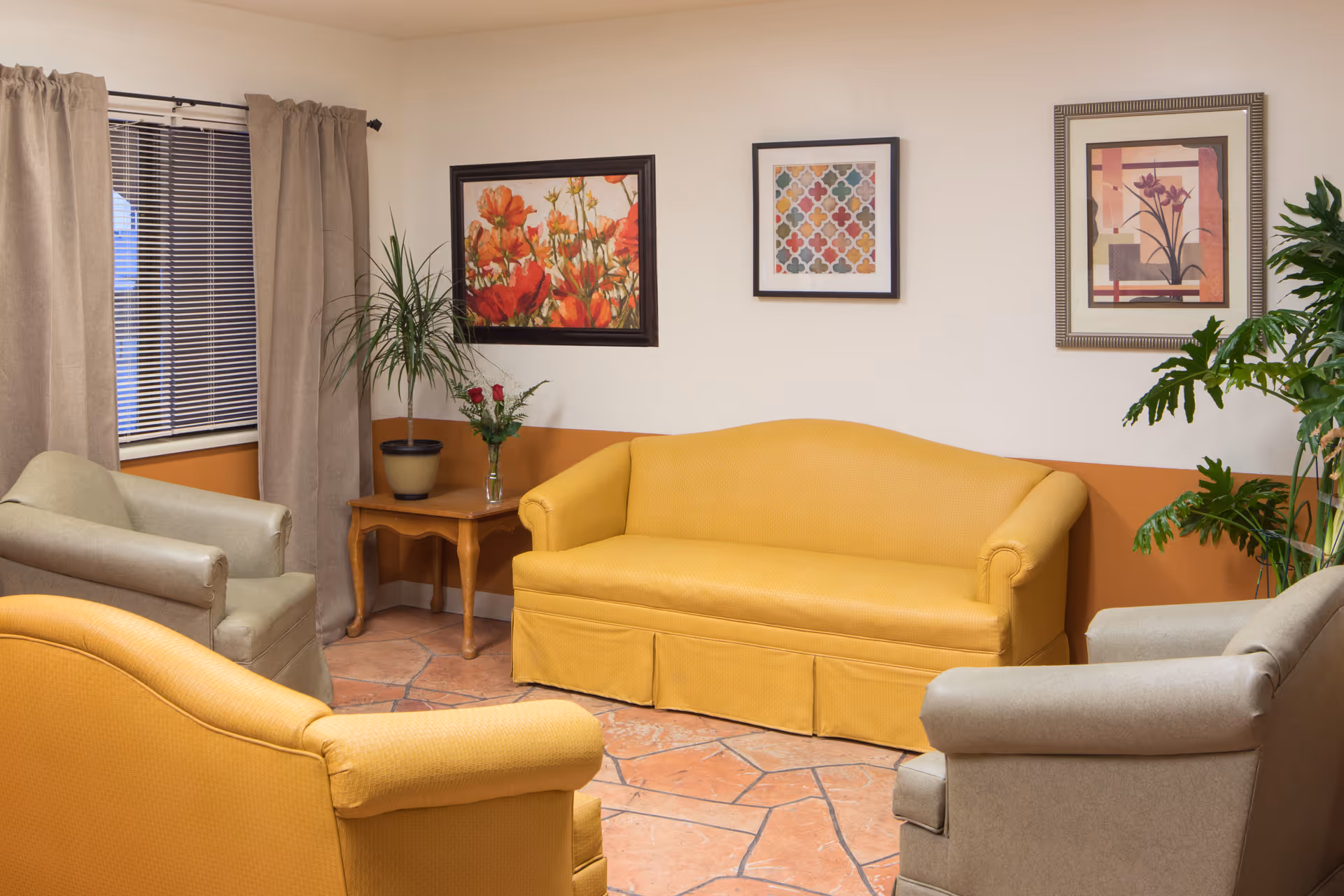 A cozy sitting area with a yellow sofa and two beige armchairs arranged around a small wooden side table holding a vase with flowers. The room has beige walls with an orange accent stripe, three framed artworks on the wall, a window with beige curtains and blinds, and two potted plants.