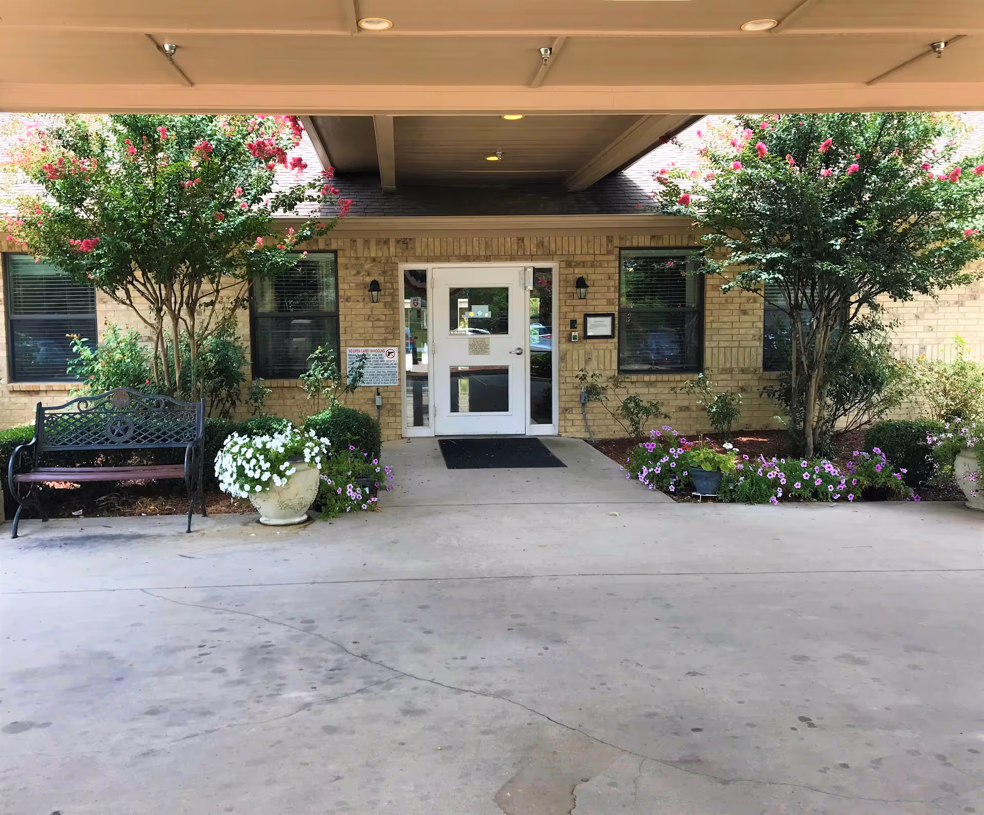 Entrance to a building with a covered driveway, featuring a white door with glass panels. There are two windows on either side of the door, surrounded by beige brick walls. The entrance is flanked by two flowering trees and various potted plants and flowers. A black metal bench is positioned on the left side near the plants.