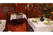 Dining room with tables covered in white tablecloths set with green plates, wooden chairs, and a china cabinet against warm red walls.