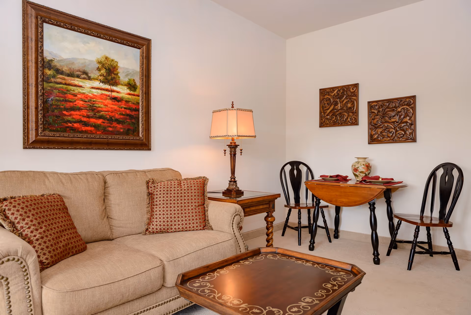 A cozy living room area featuring a beige sofa with two patterned cushions, a wooden side table with a decorative lamp, a wooden coffee table with ornate designs, and a small wooden dining table set with two chairs, a vase, and place settings. The walls are decorated with a framed landscape painting and two carved wooden wall hangings.