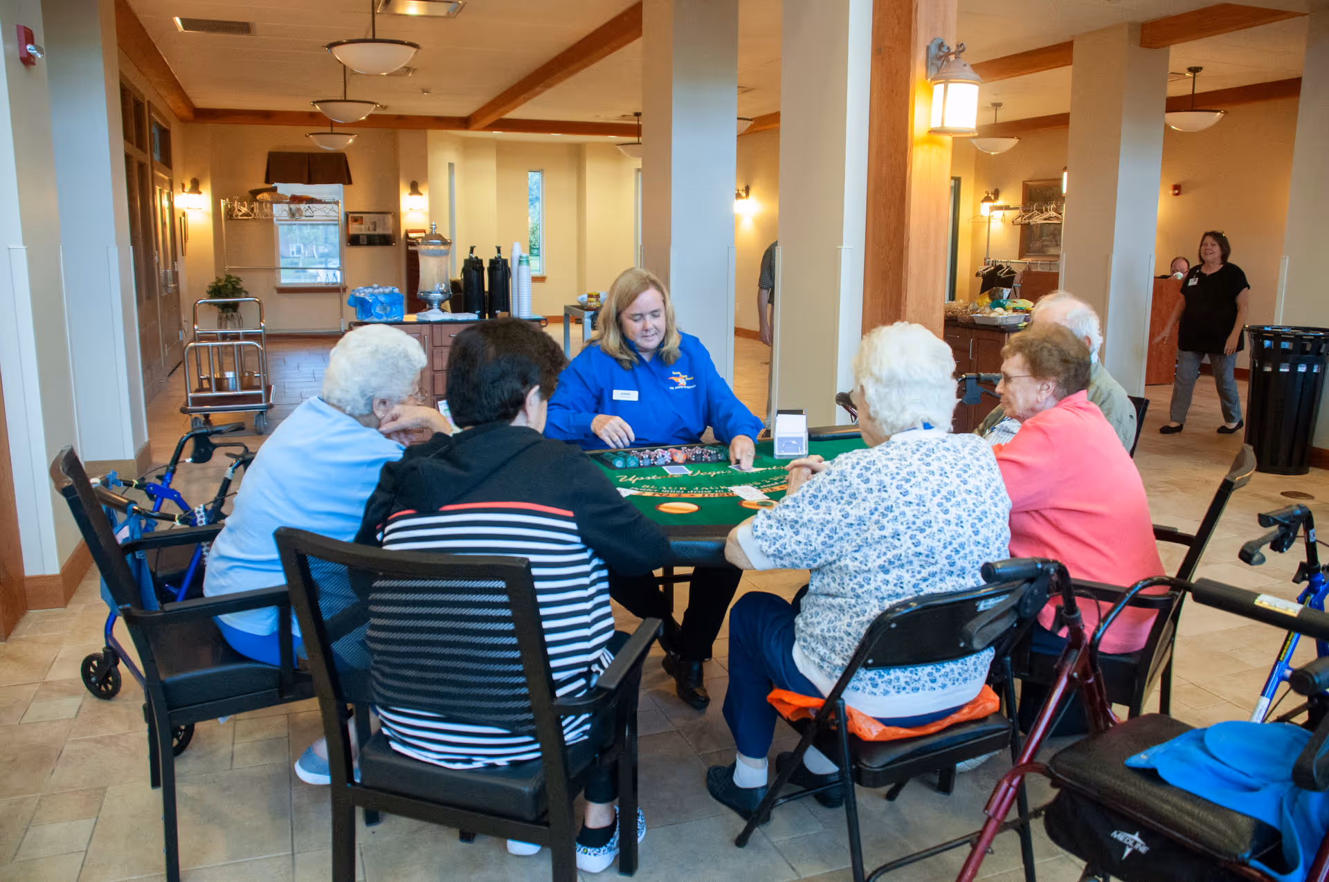 A group of elderly people sitting around a table playing a card game in a well-lit common area of a senior living facility. A staff member in a blue shirt is facilitating the game. Walkers and mobility aids are visible nearby. The room has beige walls, tiled floors, and wooden accents.