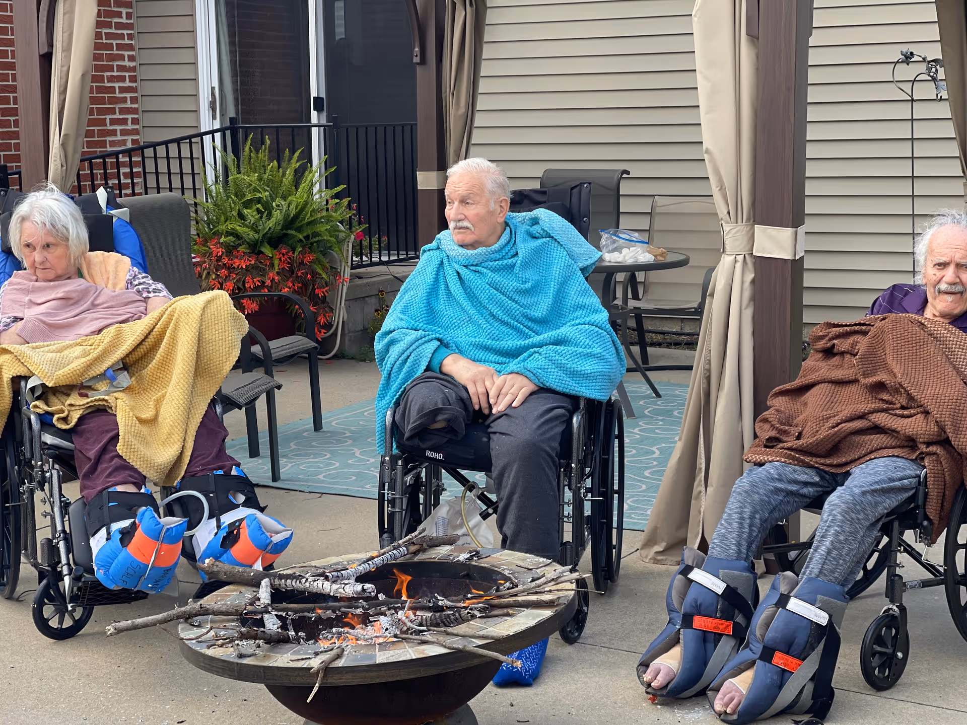 Three elderly individuals sitting in wheelchairs outdoors around a fire pit with burning logs. They are covered with blankets and wearing protective foot braces. Behind them is a building with beige siding and a patio area with plants and outdoor furniture.