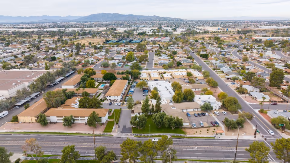 Aerial view of Tempe Post Acute facility and surrounding neighborhood, showing multiple buildings with tan roofs, parking lots with cars, trees, and streets extending into the distance with mountains visible on the horizon.