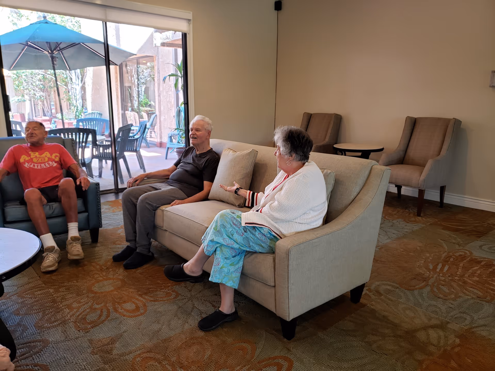 Three elderly people sitting and conversing in a comfortable living room area with beige furniture and a large window showing an outdoor patio with tables, chairs, and an umbrella.