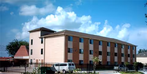 Three-story beige brick senior living facility exterior with windows, fence, and parked cars.