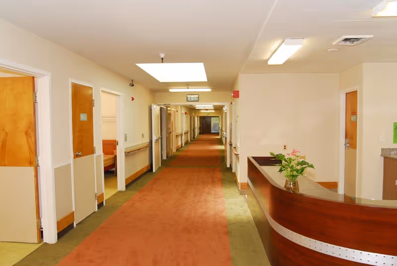 Long interior hallway of an assisted living facility with room doors lining both sides and a reception desk on the right.