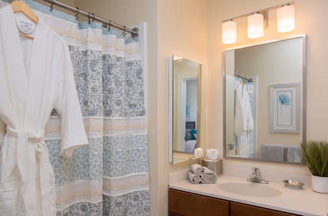 A bathroom with a white bathrobe hanging on a hook next to a shower curtain with blue and beige patterns. There is a countertop with a sink, a tissue box, rolled towels, a small plant, and a soap dish. A large mirror and a smaller mirror are mounted on the wall above the sink, reflecting part of the room.