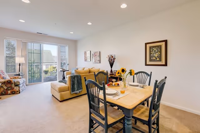 A bright living and dining area in a senior living facility with a wooden dining table set for four, decorated with sunflowers in a vase. The living room has a beige sectional sofa with cushions and a throw blanket, a floral armchair, a side table with a lamp, and large windows with a sliding glass door leading to a balcony.