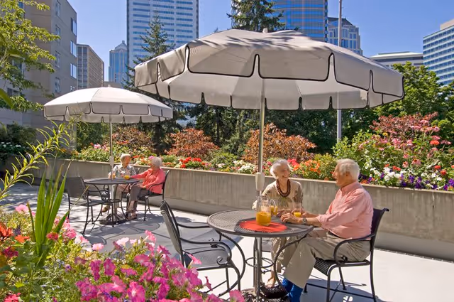 Two elderly couples sitting at separate round metal tables with umbrellas on a sunny patio surrounded by colorful flowers and greenery, with tall buildings visible in the background.