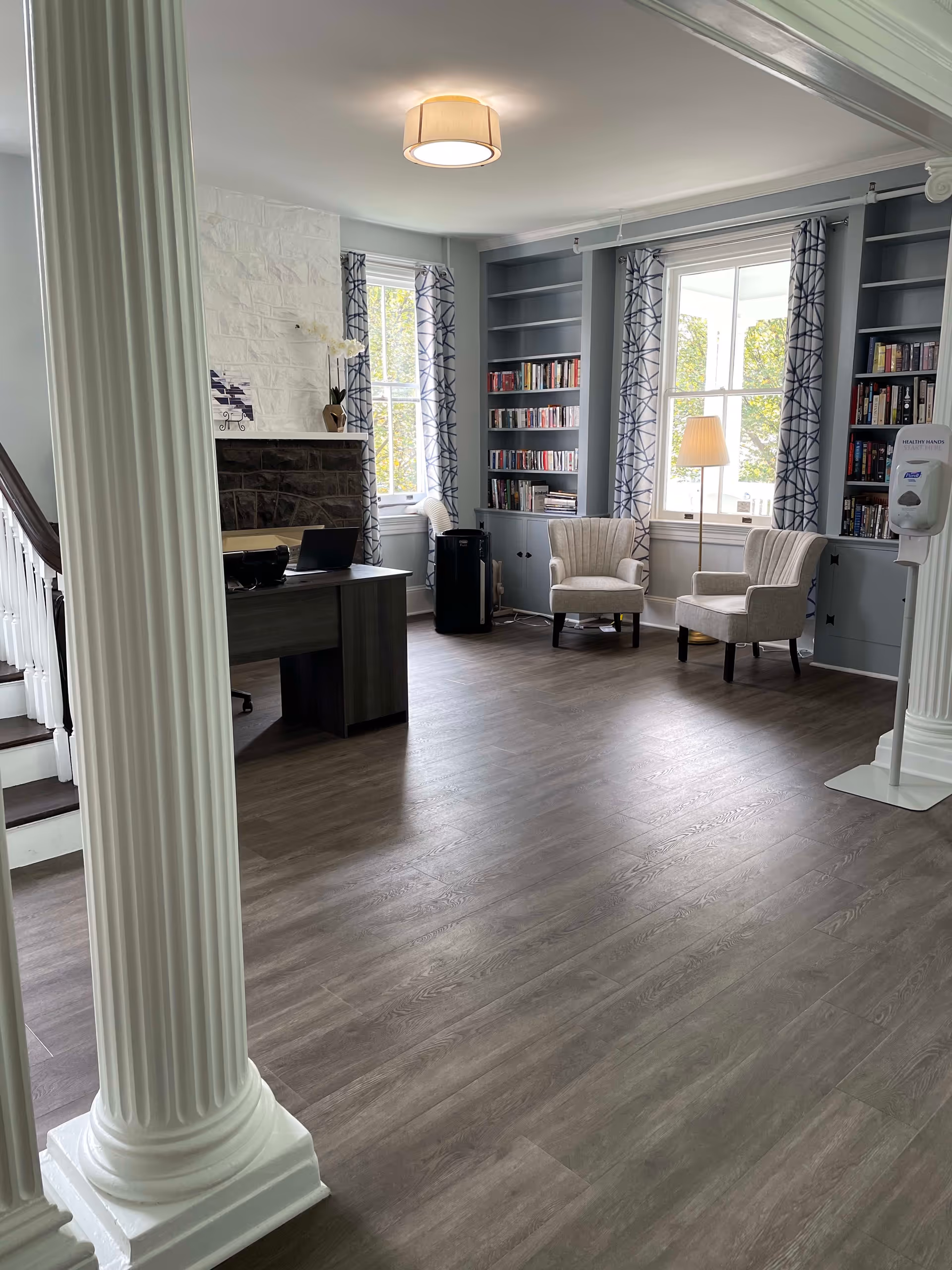A bright and spacious room with two large white columns in the foreground, a wooden desk with a laptop, two upholstered armchairs near two large windows with patterned curtains, built-in bookshelves filled with books, a floor lamp, and a hand sanitizer dispenser mounted on a stand. The room has wood flooring and a white ceiling light fixture.