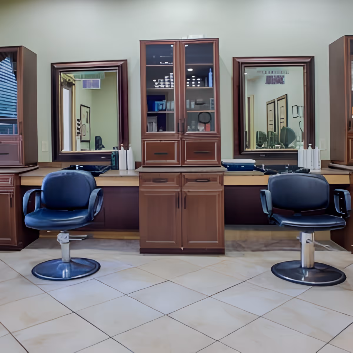 Interior view of a salon area with two black swivel chairs in front of wooden counters and large mirrors. The counters have cabinets and shelves with various items, and the floor is tiled with light-colored tiles.