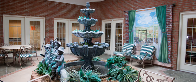 Indoor courtyard area with a multi-tiered water fountain surrounded by green plants and bronze statues of children playing. The space has brick walls, multiple glass-paneled doors, striped cushioned chairs, a table with chairs, and a large mural depicting an outdoor garden scene with green curtains painted on the wall.