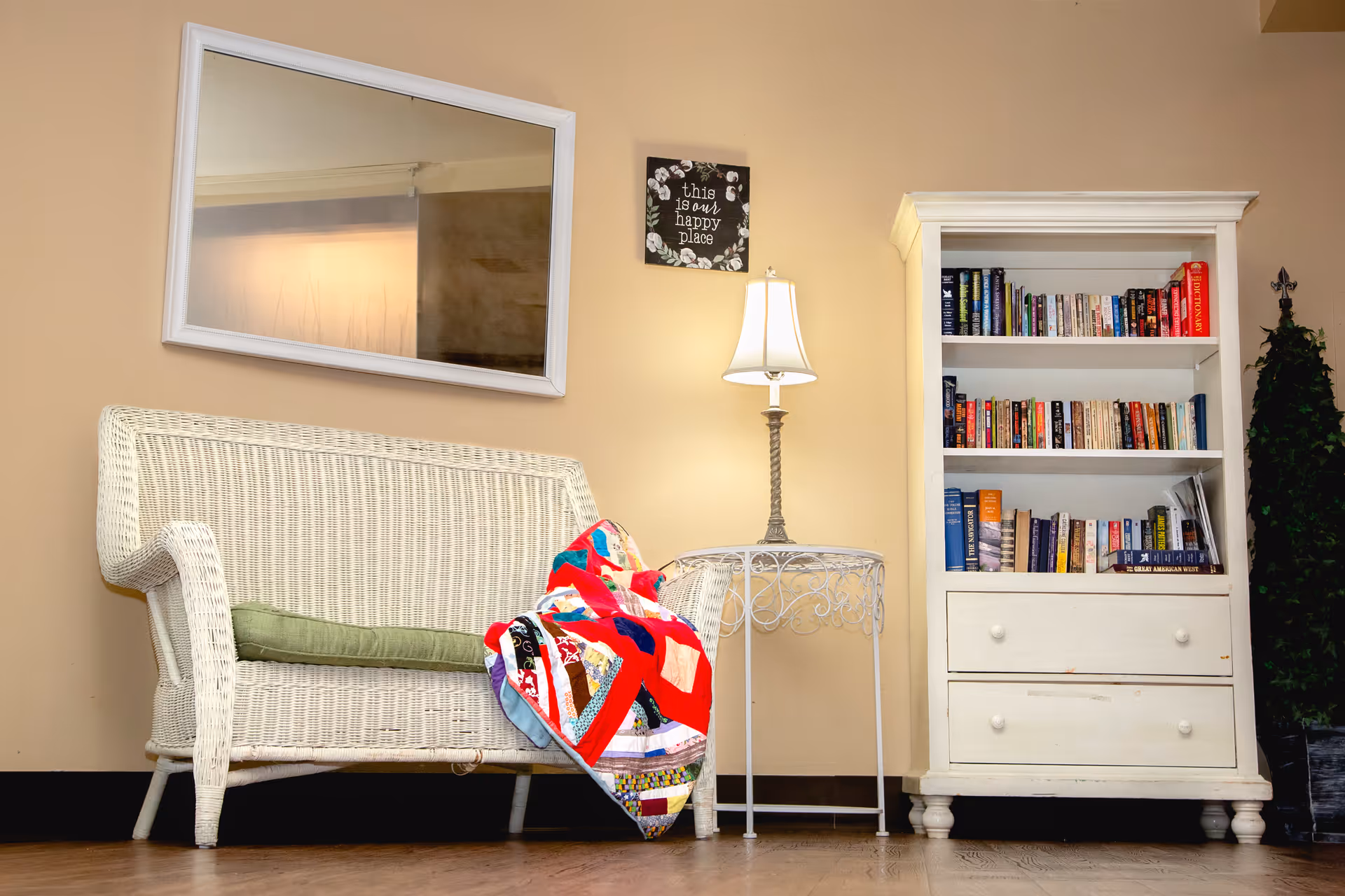 A cozy sitting area with a white wicker loveseat that has a green cushion and a colorful quilt draped over one side. Next to the loveseat is a white metal side table with a lamp on it. On the wall above the table is a small framed sign that reads 'this is my happy place.' To the right is a white bookshelf filled with books and a tall green plant beside it. The room has beige walls and wooden flooring.