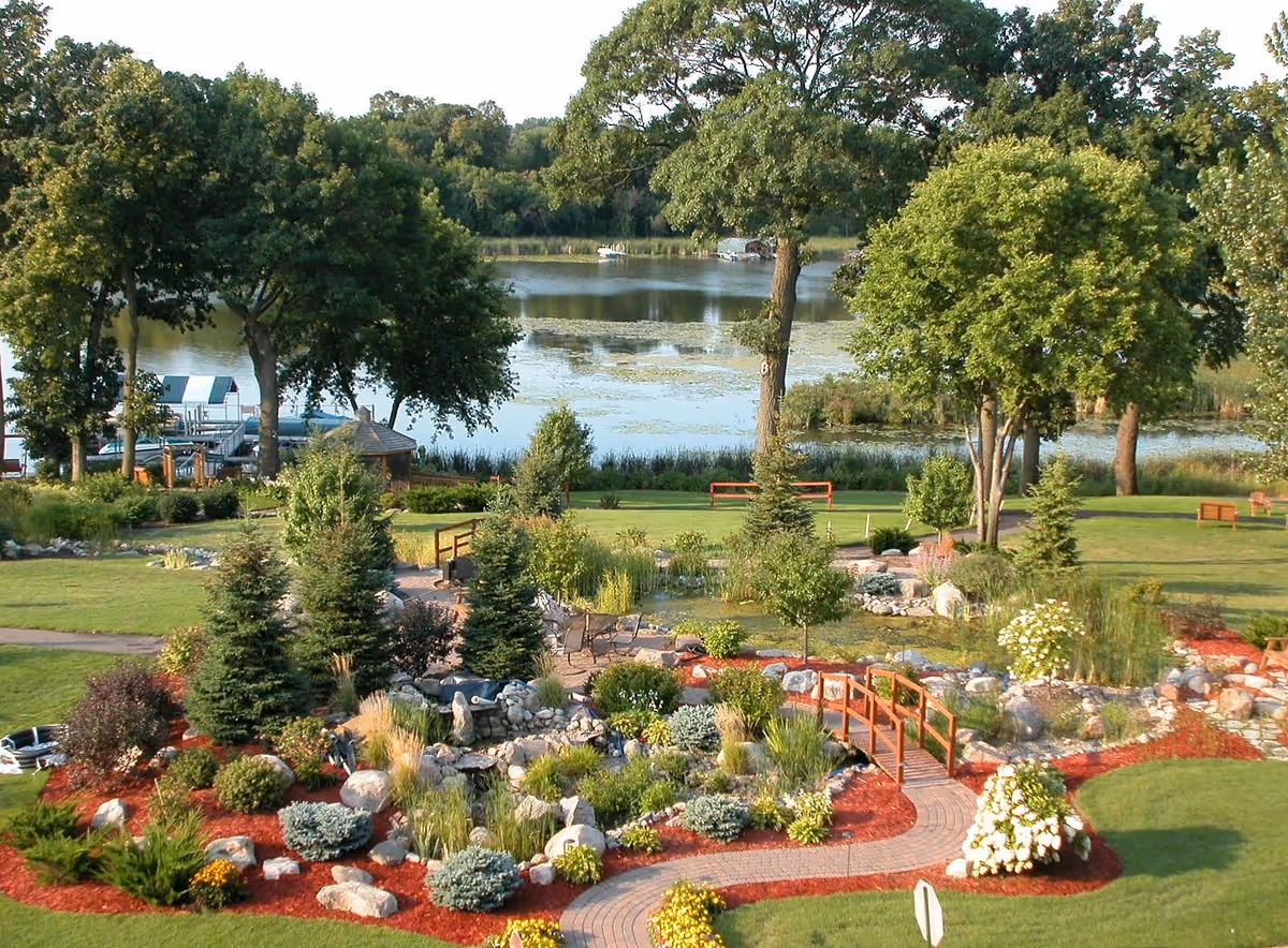 A landscaped garden area with a variety of trees, shrubs, flowers, and a small wooden bridge over a rock-lined water feature. In the background, there is a lake with boats docked and surrounded by more trees.