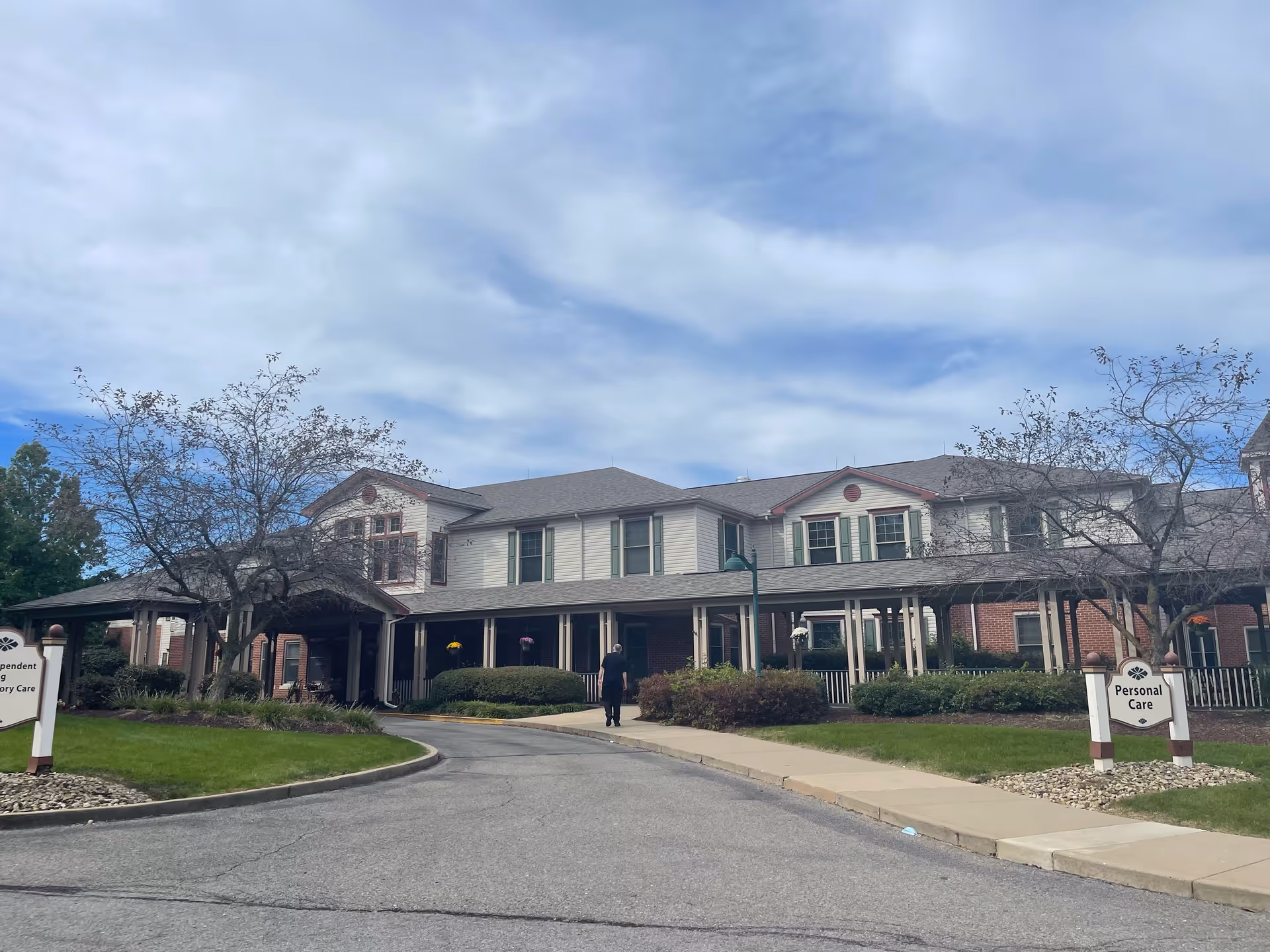 Exterior view of a senior living facility building with a covered entrance, surrounded by landscaped bushes and trees under a partly cloudy sky. A person is walking on the sidewalk towards the entrance. Signs near the driveway indicate 'Personal Care' and 'Independent Living'.