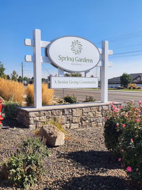 Outdoor view of a white sign for Spring Gardens Meridian, a senior living community, mounted on a stone base surrounded by landscaping with bushes, flowers, and rocks under a clear blue sky.