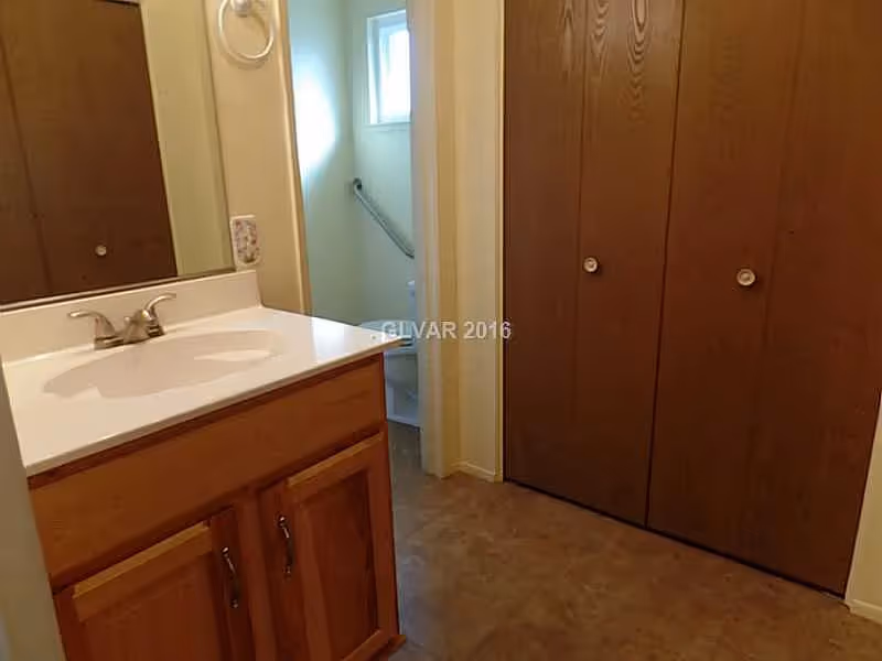 A bathroom interior featuring a wooden vanity with a white countertop and sink, a mirror above the sink, and a towel ring on the wall. In the background, there is a small window and a glimpse of a toilet with a grab bar beside it. To the right, there are large wooden closet doors.