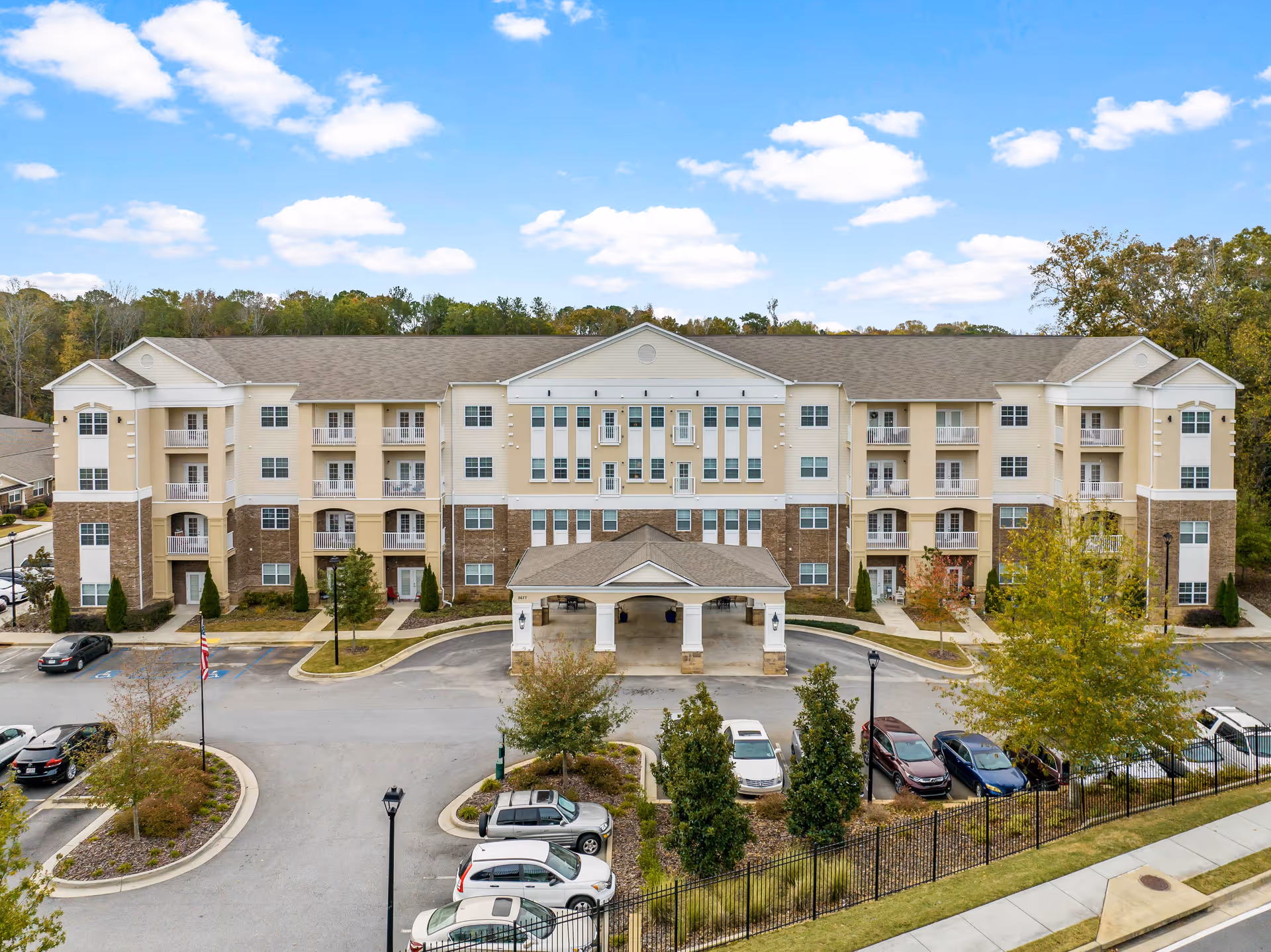 Front exterior view of a large, multi-story senior living facility building with beige and stone facade, multiple windows and balconies, a covered entrance, surrounded by parking spaces and landscaped greenery under a partly cloudy blue sky.