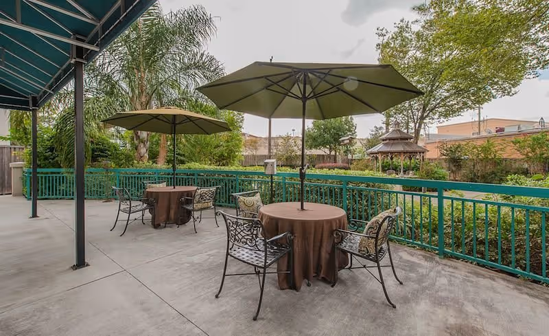 Outdoor patio area with two round tables covered with brown tablecloths, each shaded by a large umbrella. Each table is surrounded by metal chairs with decorative cushions. The patio is bordered by a green railing and overlooks a garden with trees, shrubs, and a gazebo in the background.