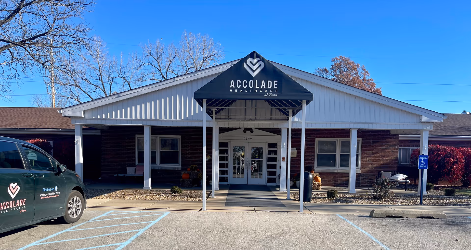 Front exterior view of a single-story brick building with a white gabled roof and a black awning that reads 'ACCOLADE HEALTHCARE of Peoria'. There is a parking lot in front with a handicap parking space and a vehicle parked on the left side with the same Accolade Healthcare logo. Trees without leaves and some red bushes are visible in the background under a clear blue sky.