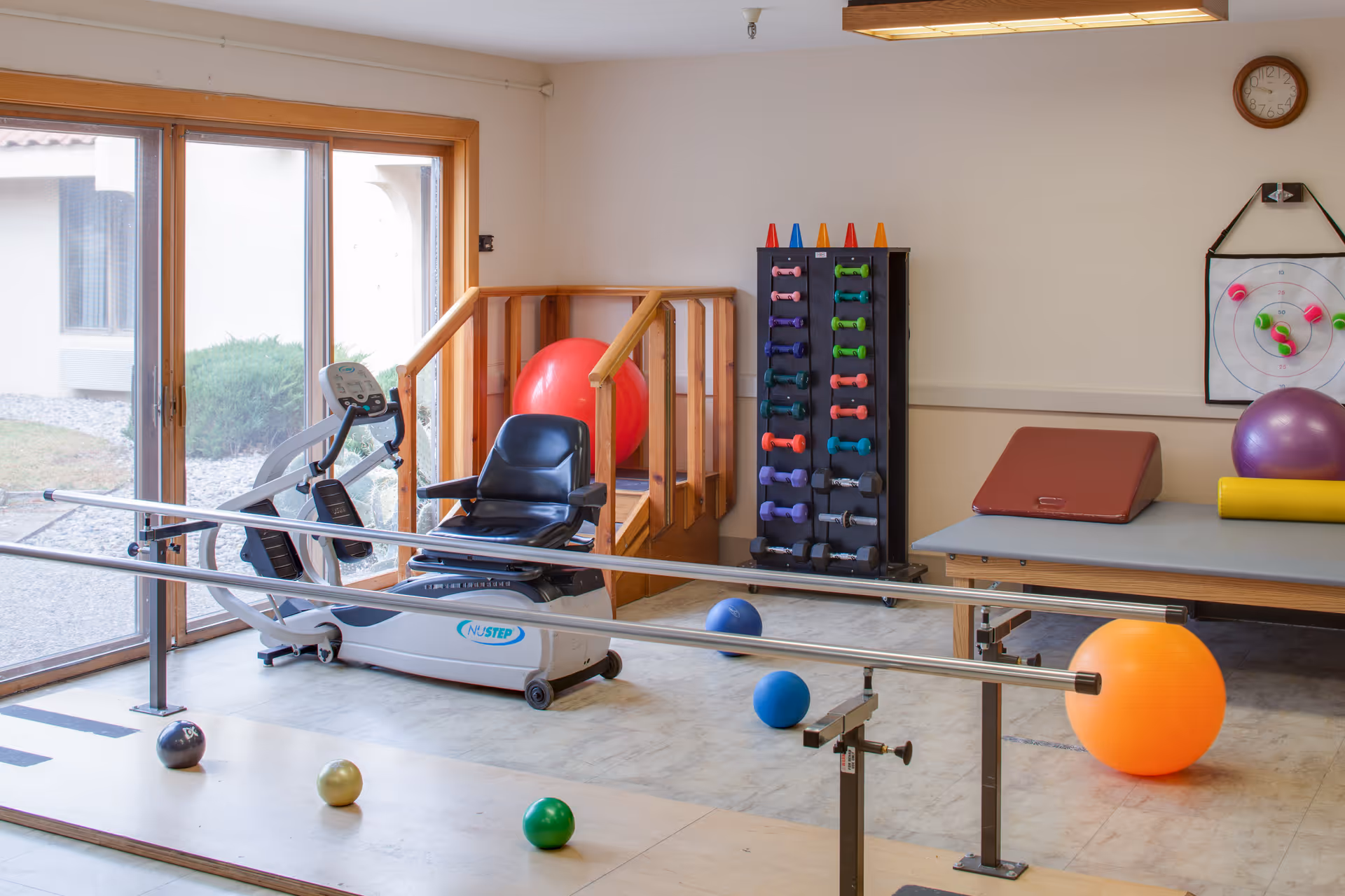 Therapy/exercise room with parallel bars, a recumbent exercise machine, stability balls, a rack of dumbbells, and a treatment table.