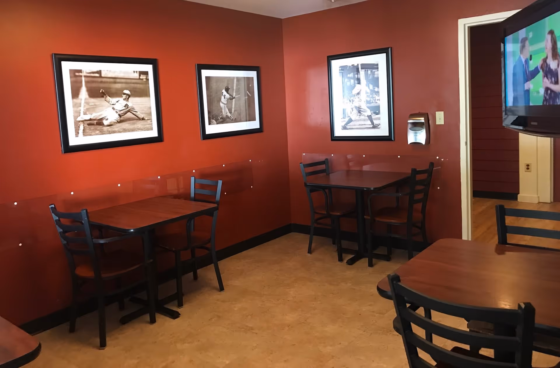 Small dining area with red walls featuring three framed black and white baseball photographs. There are three wooden tables with black chairs arranged around them. A flat-screen TV is mounted on the wall to the right, and a hand sanitizer dispenser is mounted on the wall near the doorway.