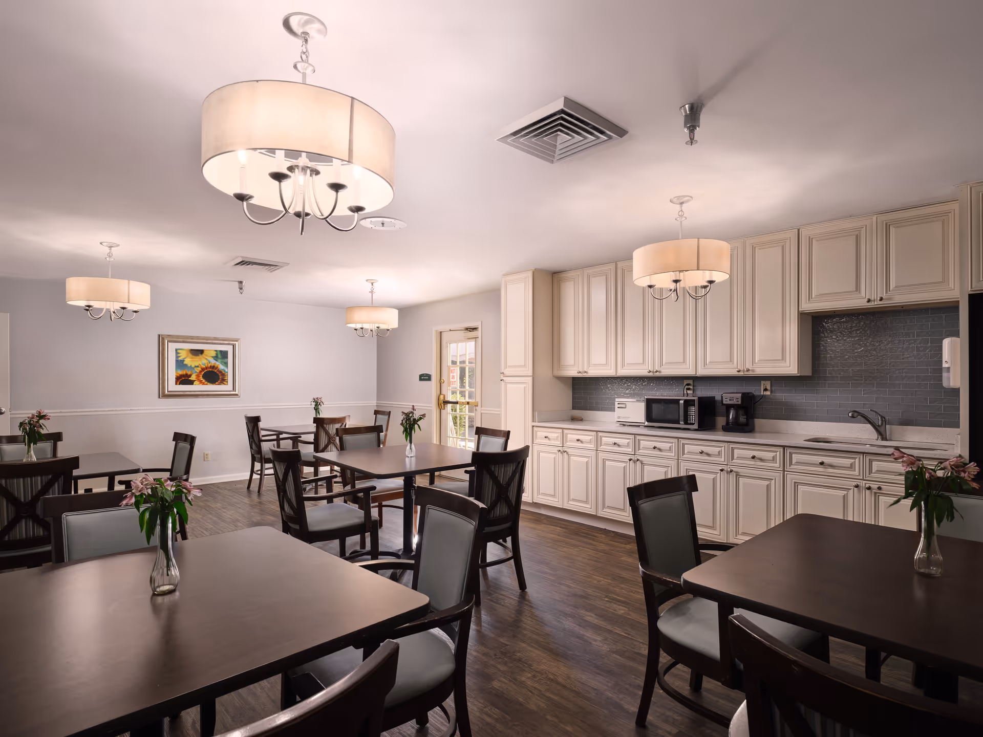 A dining room area with several dark wood tables and chairs, each table decorated with a small vase of flowers. The room features white cabinetry along one wall with a microwave, coffee maker, and toaster on the countertop. There are three large ceiling light fixtures with drum shades, wood flooring, and a framed sunflower painting on the far wall.
