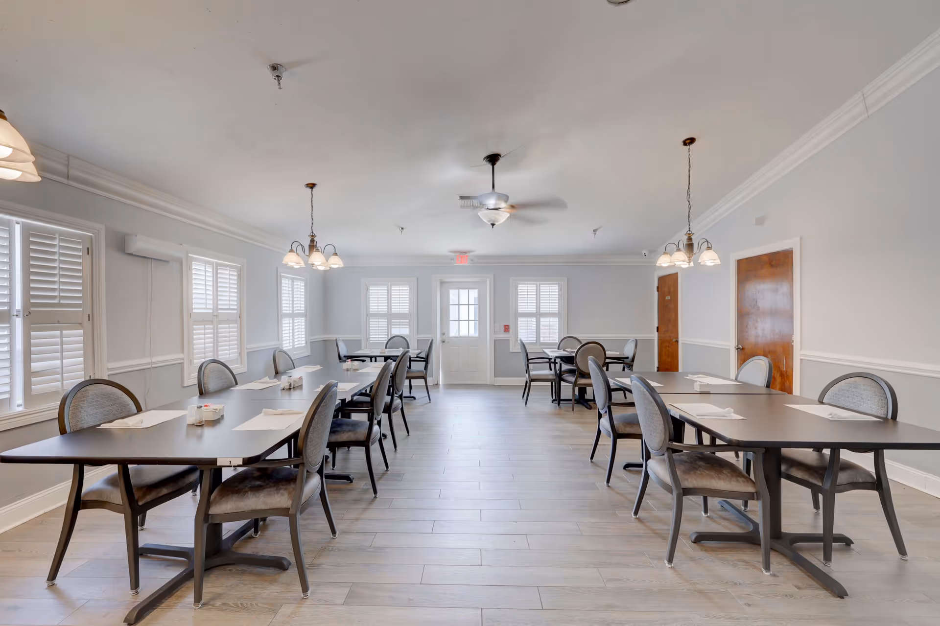 A bright and spacious dining room with multiple tables and chairs arranged neatly. The room features light gray walls, large windows with white shutters, wooden doors, and ceiling lights hanging above the tables. The floor is covered with light-colored wood planks.