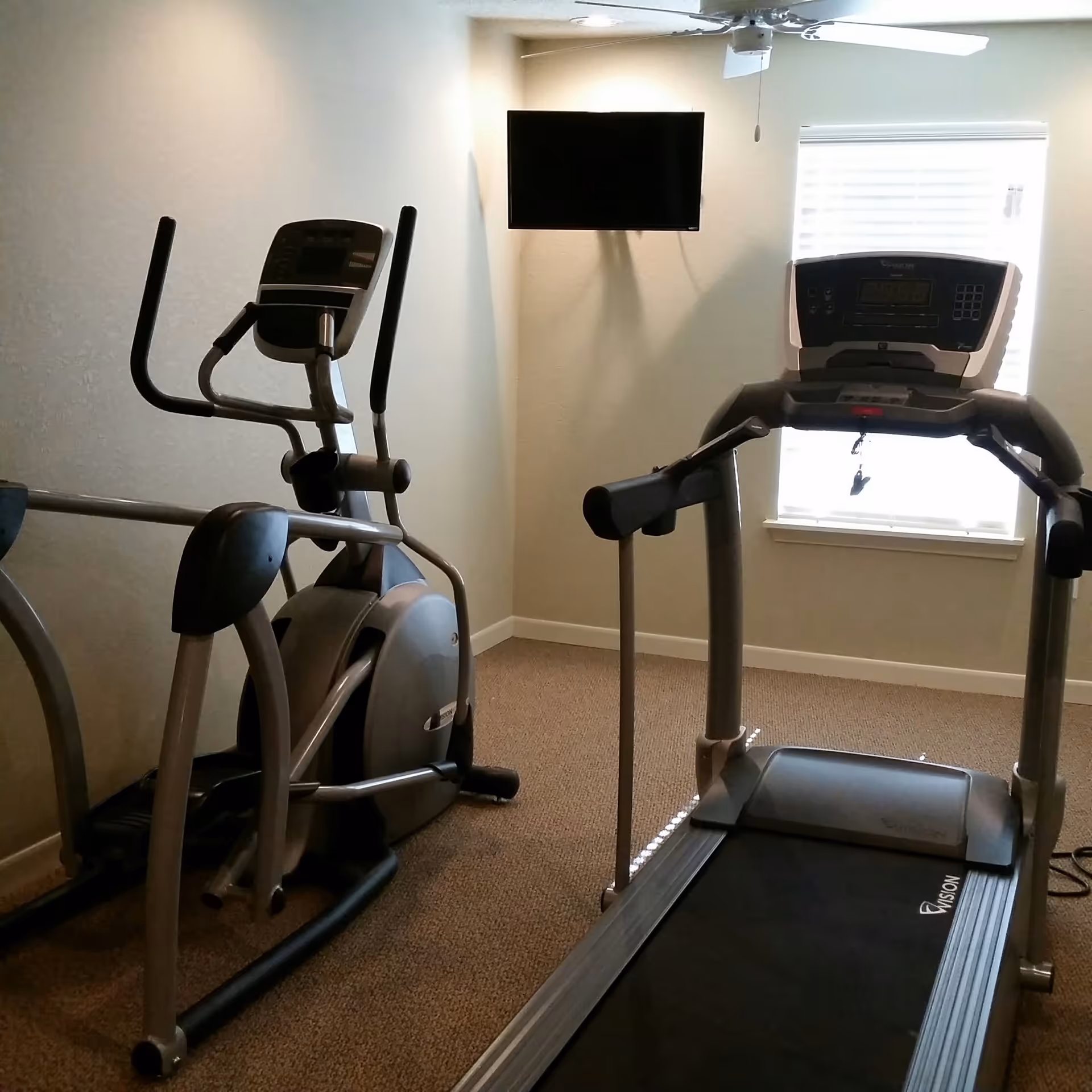 A small exercise room with a treadmill and an elliptical machine positioned on a carpeted floor. A flat-screen TV is mounted on the wall above the treadmill, and there is a window with blinds letting in natural light.