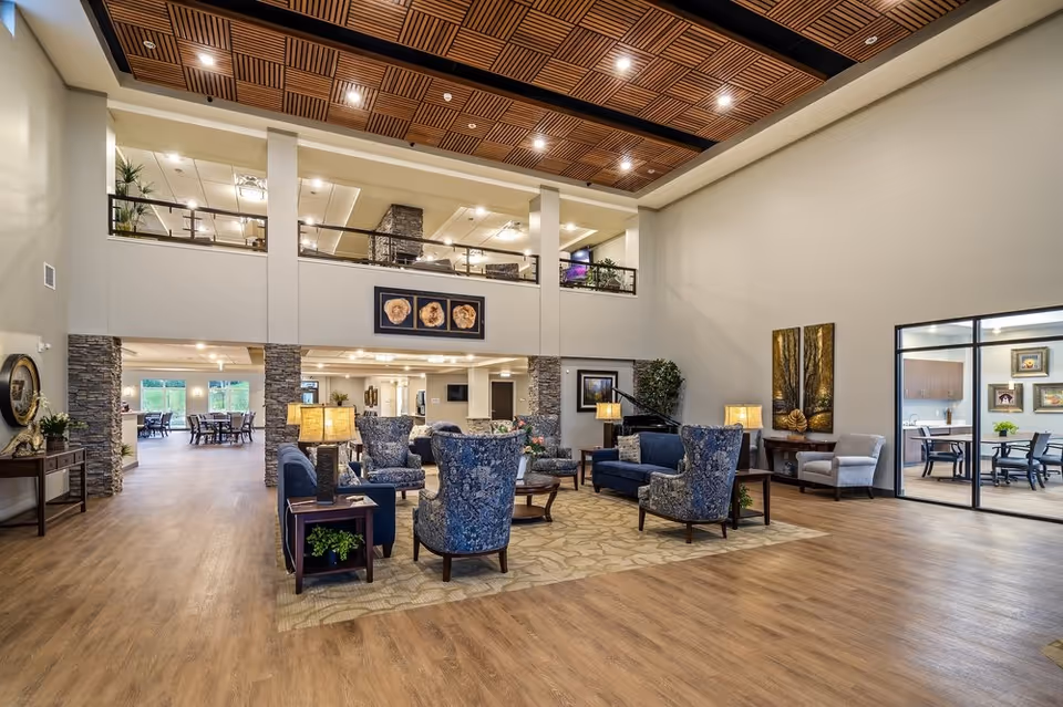 Spacious senior living facility common area with high ceilings and wood-paneled ceiling accents. The room features a seating arrangement with blue patterned armchairs, navy sofas, wooden tables with lamps, and decorative plants. In the background, there is a dining area with tables and chairs, and a glass-walled room with additional seating and framed artwork on the walls.