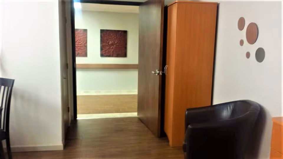 Small sitting area with a black armchair, wooden cabinet and an open door leading to a hallway with wall art.