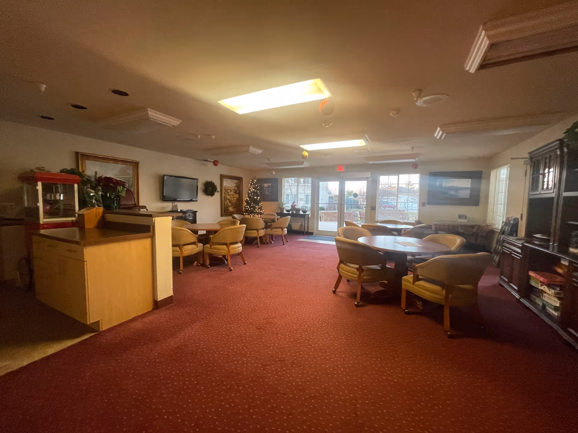 Interior view of a senior living community common area with red carpet flooring, several round tables surrounded by beige cushioned chairs, a Christmas tree decorated with lights, a popcorn machine on a wooden counter, framed paintings on the walls, a flat-screen TV mounted on the wall, and large windows and glass doors letting in natural light.